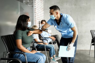 Arap genç doktor, kadın hasta Afro-Amerikan bayanı dirsek darbesiyle karşılıyor. Klinikte oturan, COVID-19 'a karşı aşı bekleyen, yüz maskesi takan çok ırkçı bir grup insan.