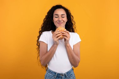 Mutlu bir bayan model stüdyo ortamında lezzetli bir burgeri tutarken ve koklarken gülümser. İştah açıcı fast food 'tan bir ısırık almaya hazırlanırken heyecanını gösteriyor..