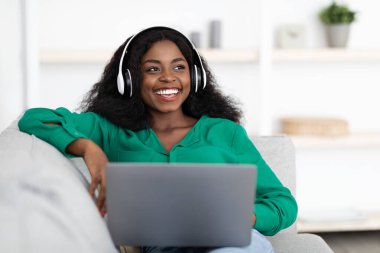 Bir kadın kanepede rahat bir şekilde otururken dizüstü bilgisayarında müzik dinlemekten zevk alır. Rahatlamış, kulaklık takıyor ve şık bir evde gülümsüyor..