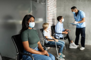 COVID-19 'a karşı aşı bekleyen uluslararası erkek ve kadın hastalar, hastane koridorunda oturan erkek doktor Afro-Amerikan kadın maskeli kadınla konuşuyorlar.