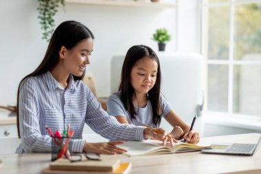 Parlak bir mutfakta, genç bir Asyalı anne kızına ev ödevinde yardım eder. Mutlu kız bir deftere yazarken annesi önündeki bir şeyi işaret ediyor ve öğrenmesini teşvik ediyor..