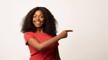 Neşeli bir Afro-Amerikan kadın gülümsüyor ve parmağıyla işaret ediyor. Sade beyaz bir arka plana karşı poz veriyor, promosyon içeriği için hoş bir ortam yaratıyor..
