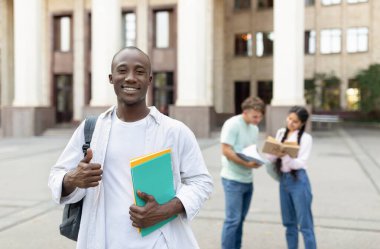 Bir üniversite öğrencisi renkli klasörler tutarken kendine güvenerek başparmağını kaldırıyor. Arka planda, iki sınıf arkadaşı bir üniversitenin dışında eğitim görüyorlar..