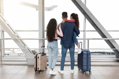 Üç kişilik aile havaalanında, havalanan bir uçağa büyük pencereden bakıyorlar. Bavullarının yanında duruyorlar, yolculukları için heyecanlılar..