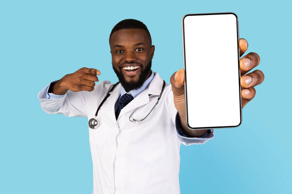 Excited African American Doctor Showing Smartphone With Blank Screen At Camera While Standing Over Blue Background, Black Medical Worker Pointing At Mobile Phone Screen, Recommending App, Mockup