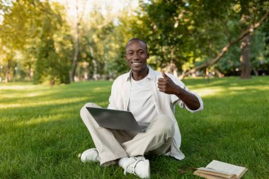 Güneşli bir parkta, genç bir adam dizüstü bilgisayarıyla çimlerin üzerinde oturuyor, gülümsüyor ve başparmağını kaldırıyor. Etrafı doğa ile çevrili rahatlatıcı bir andan hoşlanıyor, yakınlarda bir kitap okuyor..