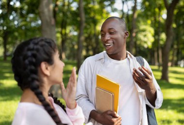 İki arkadaş ağaçlarla dolu yeşil bir parkta canlı bir sohbetin tadını çıkarıyorlar. Ellerinde çalışma materyalleri varken gülümsüyorlar ve mutlu görünüyorlar..
