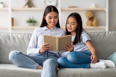 Genç bir Asyalı anne ve kızı rahat bir kanepede oturup birlikte kitap okuyorlar. Rahatlatıcı bir ortamda, evde mutlu bir anı paylaşırlar, gülümsemelerini paylaşırlar..