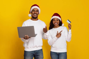 Noel Baba şapkası takan neşeli bir çift parlak sarı bir arka plana karşı duruyor. Sırıtıyorlar ve ellerinde bir dizüstü bilgisayar ve kredi kartı tutarken online alışveriş hakkında heyecan gösteriyorlar..