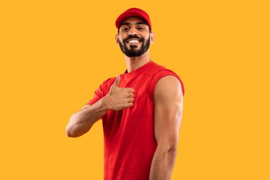 Kırmızı kısa kollu gömlek giyen genç bir adam başparmak işareti gösterirken geniş bir gülümseme sergiliyor. Kolunda küçük bir bandaj var. Aşı olduğunu gösteriyor..