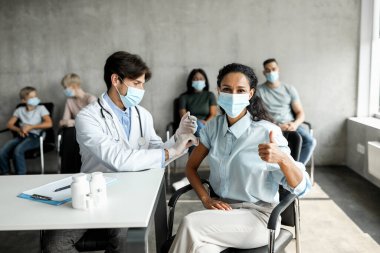 Orta doğulu kadın hasta klinikte koronavirüse karşı aşı oluyor, kameraya baş parmağını gösteriyor, genç doktor esmer bir kadın için omuzdan kaslar arası çekim yapıyor.