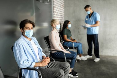 Klinikte oturan, koronavirüse karşı aşı bekleyen koruyucu maskeli çok ırklı hastalar, genç yakışıklı Arap doktorla sohbet eden, aşılama konsepti