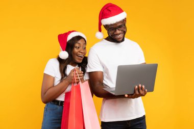 Noel Baba şapkası takan neşeli siyah bir çift online Noel alışverişi yapıyor. Renkli alışveriş çantaları tutuyorlar ve canlı sarı bir arkaplanda dizüstü bilgisayar kullanırken gülümsüyorlar..