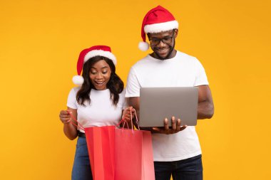 Neşeli bir siyah çift, Noel Baba şapkası takarken dizüstü bilgisayar kullanarak internetten alışveriş yapıyor. Renkli alışveriş torbaları taşıyorlar, canlı sarı bir arka plana karşı Noel sezonunu kutluyorlar..