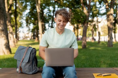 Genç bir adam parktaki ahşap bir bankta otururken dizüstü bilgisayarına odaklanarak gülümsüyor. Çevreleyen ağaçlar ve yeşil otlar huzurlu bir atmosfer yaratıyor..