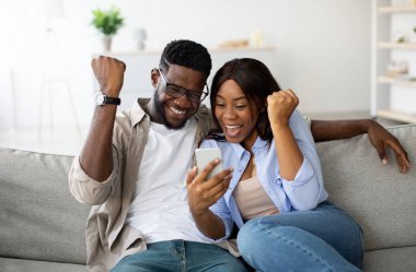 Afro-Amerikalı bir çift koltukta oturup akıllı telefonlarından harika haberler veriyor. Online galibiyetlerini kutlamak için yumruklarını kaldırıyorlar. Saf neşe ve beraberlik sergiliyorlar..