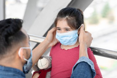 Bir baba, küçük kızına oyuncak ayısını tutarken maske takması için nazikçe yardım eder. İçerideler, muhtemelen halka açık bir yerde, gündüz vakti..