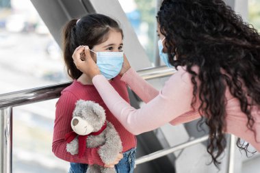 Bir anne genç kızına maske takmasında nazikçe yardım eder. Kız bir oyuncak ayı tutuyor, ve onlar parlak, çağdaş kapalı bir alanda, güvenlik ve bakım üzerine odaklanıyorlar..