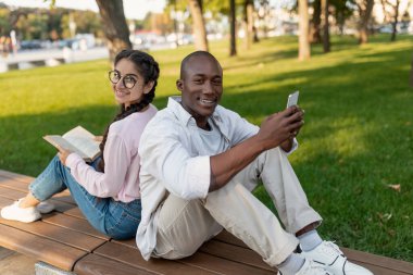 İki arkadaş arka arkaya parkta bir bankta oturur, biri kitap okurken diğeri akıllı telefon kullanır. Ağaçlar ve otlar öğleden sonra güneşinde huzurlu bir ortam yaratır..