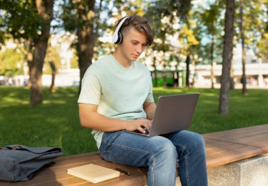 Parktaki bankta oturan genç bir adam kulaklık takarken dizüstü bilgisayarına odaklanmış. Ağaçların arasında güneşli bir öğleden sonra geçirir, iş ve dinlenmeyi dengeler..