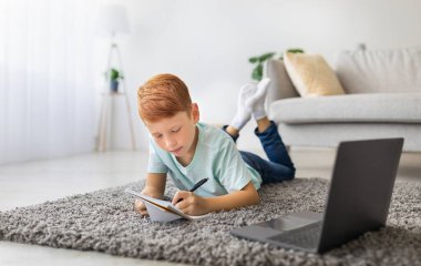 Kızıl çocuk halıya rahatça uzanır, dizüstü bilgisayar kullanırken not almaya odaklanır. Ev ödevi yapıyor ya da sıcak bir oturma odasında online bir derse katılıyor..