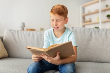 Sevimli kızıl saçlı bir çocuk evdeki rahat bir kanepede oturuyor, elinde kitap tutuyor. Okurken gülümsüyor, içindeki hikayelere ve maceralara derinden bağlanıyor. Huzurlu bir odak ve sevinç anı.