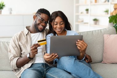 Parlak bir oturma odasında, genç bir Afrikalı Amerikalı çift online alışveriş yapıyor. Kadın elinde tabletle gülümserken, adam kredi kartıyla çıkış yapıyor. İkisi de satın almaları konusunda heyecanlı..