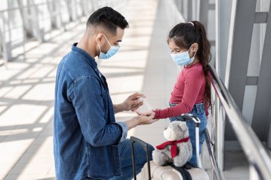 Bir baba, havaalanında beklerken kızına bavulunda yardım eder. İkisi de maske takıyor ve bagaja oyuncak ayı oturuyor..