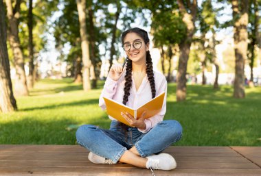 Gözlüklü genç bir kız parkta tahta güvertede bacak bacak üstüne atmış oturuyor. Elinde bir defter tutarken gülümsüyor, ağaçlar ve çimenler arasında güneşli günün tadını çıkarıyor..