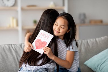 Sevgi dolu bir kız, anneler gününü annesini kucaklayarak kutlar. Evdeki rahat kanepede oturup özel anlarının tadını çıkarırken elinde bir kalp ile bir tebrik kartı tutuyor..