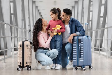 Havaalanında bir aile bir arada duruyor, gülümsüyor ve birlikte eğleniyorlar. Genç bir kız, ailesi yanında bavullarıyla diz çökerken oyuncak ayıya sarılıyor..