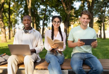 Parktaki bir bankta oturan üç öğrenci, cihazlarına odaklanmış. Ödevlerini yaparken güneş ışıl ışıl parlıyor. Sıcak havanın tadını çıkarıyorlar..