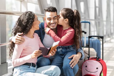 Mutlu bir aile havaalanında birlikte oturur, gülümser ve anı paylaşır. Babası, kızı onu öperken seyahat belgelerini elinde tutuyor ve annesi sevinçle bakıyor..