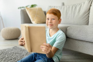 Neşeli kızıl saçlı bir çocuk yerde oturup evde kitap okuyor. Kameraya gülümsüyor, büyüleyici maceralarının heyecan verici hikayelerine dalmış, rahat bir çevreyle çevrili..