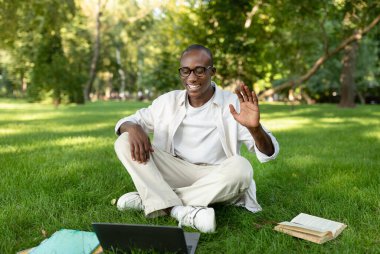 Genç bir adam parkta çimlerin üzerinde oturuyor, bir online toplantı sırasında gülümsüyor ve dizüstü bilgisayarına el sallıyor. Yakınlarda bir kitap ve sırt çantası var, dışarıda rahat bir çalışma alanının keyfini çıkardığını gösteriyor..