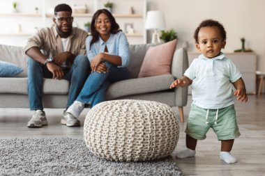 Neşeli bir bebek ayakta duruyor ve sıcak bir oturma odasında ilk adımlarını atıyor. Ailesi kanepede oturmuş, yürümeyi keşfederken ona gülümsüyor ve destekliyor..