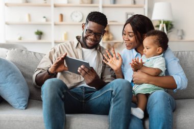 Gülümseyen bir Afro-Amerikalı erkek, kadın ve çocuk oturma odalarındaki bir kanepede oturup bir video görüşmesi sırasında tablete el sallıyorlar. Sevdikleriyle anlarını paylaşıyorlar, birlikte neşeli anılar yaratıyorlar..