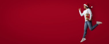 Noel Baba şapkası takan biri, elinde parlak kırmızı bir arkaplanda hediye tutarken mutlu bir şekilde zıplar. Sahne, tatil sezonunun neşesini ve şenlik ruhunu yakalıyor..