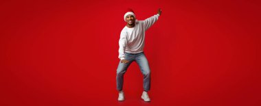Şenlikli bir süveter giyen ve Noel Baba şapkası takan bir adam canlı kırmızı bir arka planda mutlu bir şekilde dans ediyor. Neşeli ifadesi kutlama ruhunu ve tatil neşesini yansıtıyor..