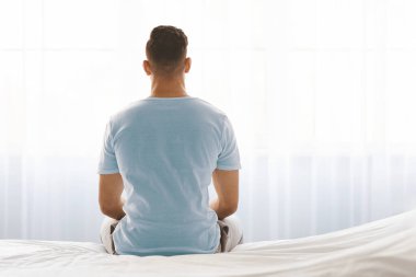 Bir adam yatağında bağdaş kurmuş, parlak bir pencereye bakıyor. Yumuşak sabah ışığı perdelerin arasından süzülerek yansıma ve meditasyon için barışçıl bir atmosfer yaratır..