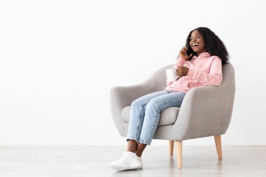 Neşeli bir kadın yumuşak bir sandalyede otururken telefonla konuşmaktan zevk alır. Elinde bir bardak tutuyor ve rahat giyiniyor. Sıcak ve davetkar bir ortam yaratıyor..