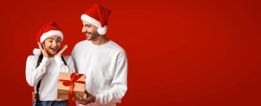 Neşeli bir çift, parlak kırmızı arka planda neşeli şapkalar takıyor. Bir anlık heyecanı paylaşıyorlar. Güzelce paketlenmiş, ışıldayan bir bayram ruhuna hayran oluyorlar..