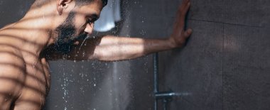 Bir adam duşun altında duruyor, sıcak suyun üzerine akmasına izin veriyor. Yumuşak ışık duvarda gölgeler oluşturur ve banyonun huzurlu atmosferini artırır..