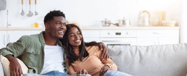 Genç bir çift, güneşli bir salonda rahat bir koltukta oturup gülüyor ve gülümsüyor. Mekan yumuşak mobilyalar ve parlak dekorla sıcak, rahat bir atmosfer yaratıyor..