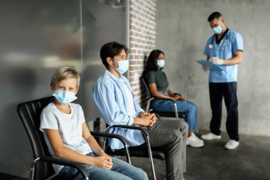 Koruyucu yüz maskesi takmış genç bir çocuk COVID-19 'a karşı aşı bekliyor. Çok ırklı bir grup hasta klinikte aşılanmaya hazırlanıyor. Ortadoğu doktoruyla sohbet ediyor.