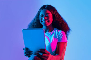 Uzun, kıvırcık saçlı genç bir kadın elinde tuttuğu tablete gülümsüyor. Arka planda parlak mavi ve pembe ışık var, canlı bir atmosfer yaratıyor..