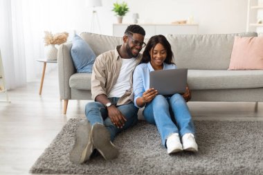 Genç Afrikalı Amerikalı çift oturma odalarının zemininde bir kanepeye yaslanarak oturuyorlar. Birlikte video izlemek ya da görüntülere göz atmak için bir dizüstü bilgisayar kullanıyorlar..