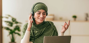 Konforlu bir ofiste, yeşil atkı ve kulaklık takan genç bir kadın bilgisayarına konuşurken gülümsüyor. Bitkilerle çevrili sanal bir konuşma sırasında meşgul ve mutlu görünüyor..