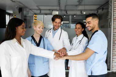 Birbirlerini motive eden çok ırklı doktorlardan oluşan bir ekip sabahları brifing yaparken, ellerini birbirinin üstüne koyup gülümserken, modern klinik iç mekanlarında. Takım çalışması, profesyonel takım konsepti.