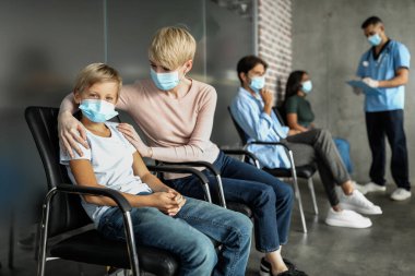 Sevgi dolu anne sarışın kadın, klinikte Coronavirus aşısı beklerken oğlunu teselli ediyor. Sırada bekleyen çok ırklı hastalar, doktorla sohbet ediyorlar.